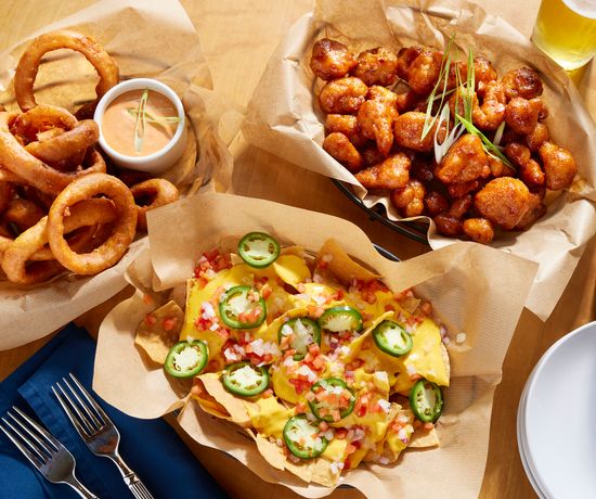 Three baskets of bar food: onion rings with dipping sauce, spicy coated cauliflower bites garnished with scallions, and nachos topped with cheese, jalapeños, tomatoes, and onions. A glass of beer and a can are nearby.