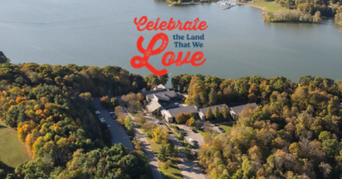 Aerial view of Mohican Lodge nestled among colorful autumn trees along the shoreline of a large lake. The image features the slogan "Celebrate the Land That We Love" above the lodge, highlighting the property's scenic natural setting and surrounding forest landscape.