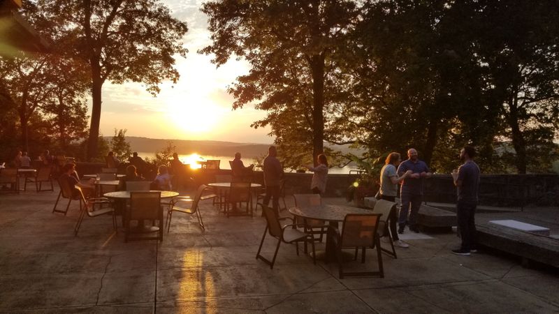 Guests gather on the outdoor terrace at Mohican Lodge during sunset, enjoying conversation and refreshments beneath towering trees as golden light fills the scenic overlook.