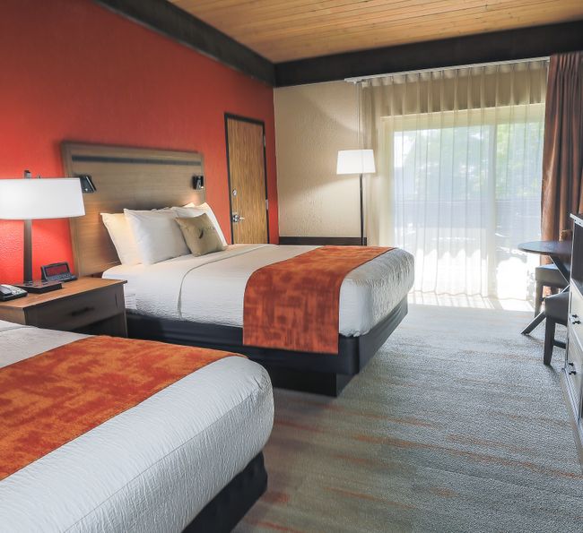 Guest room with two queen beds, warm wood accents, bedside lighting, desk and chair, flat-screen TV, and large window overlooking outdoor scenery.