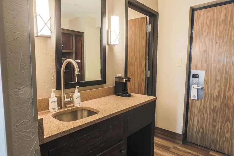 Guest room vanity area featuring a granite countertop with a sink, large framed mirror, modern wall sconces, coffee maker, and wood-accented entry door, showcasing the updated accommodations at Mohican Lodge.